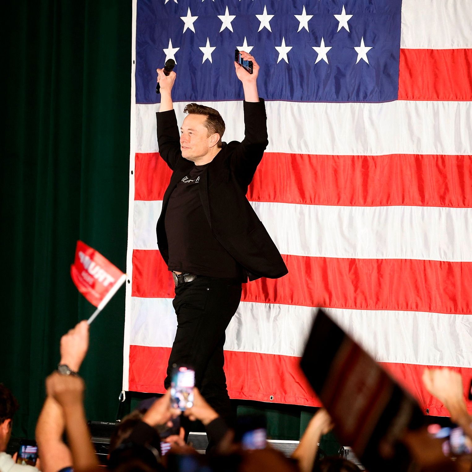 马斯克with his arms in the air as he acknowdges the crowds while standing on a stage with an enormous US flag behind him